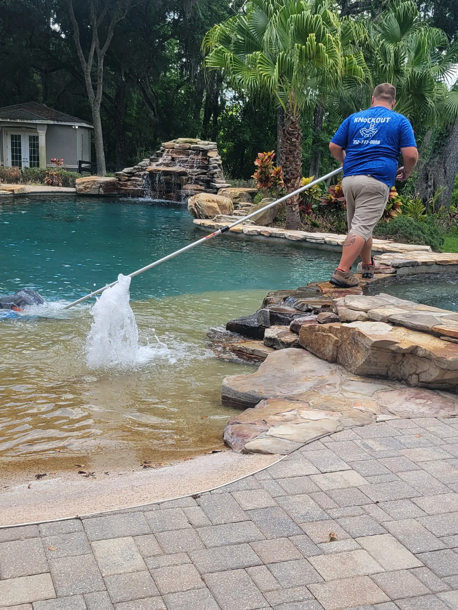 Brent from Knockout Pool & Spa Care brushing a beach-entry pool with a rock waterfall